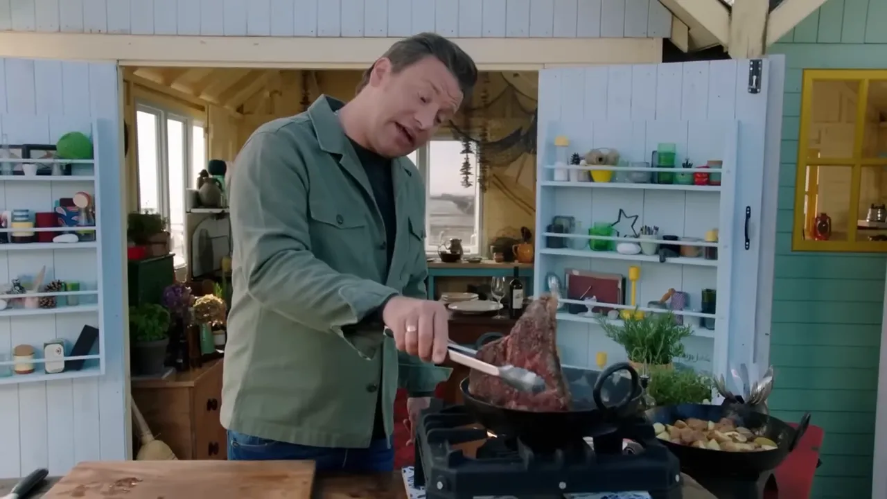 Thick T-bone steak being seared in a pan on a stovetop with a pan of roast potatoes to the side