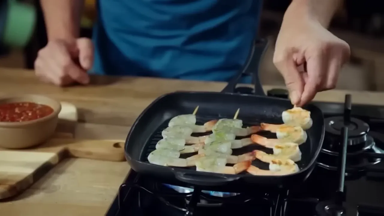 Skewered prawns cooking in a ridged pan with a small bowl of sweet chili sauce and a cutting board nearby