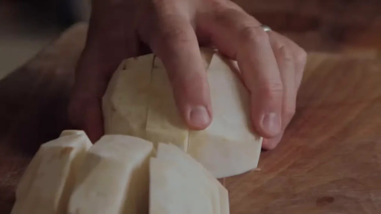 Close-up of celeriac cut into uniform chunks on a wooden chopping board with a hand steadying the root