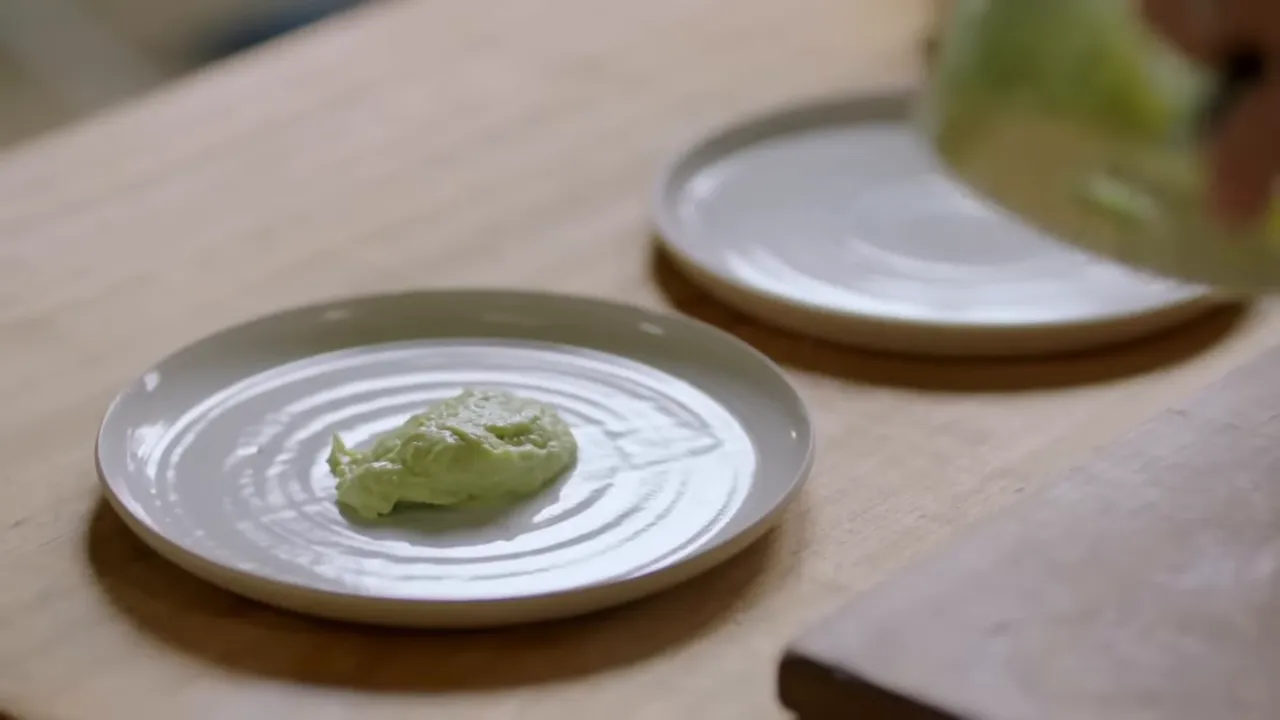 silky green smashed avocado dollop on a white plate on a wooden work surface, with another plate in the background
