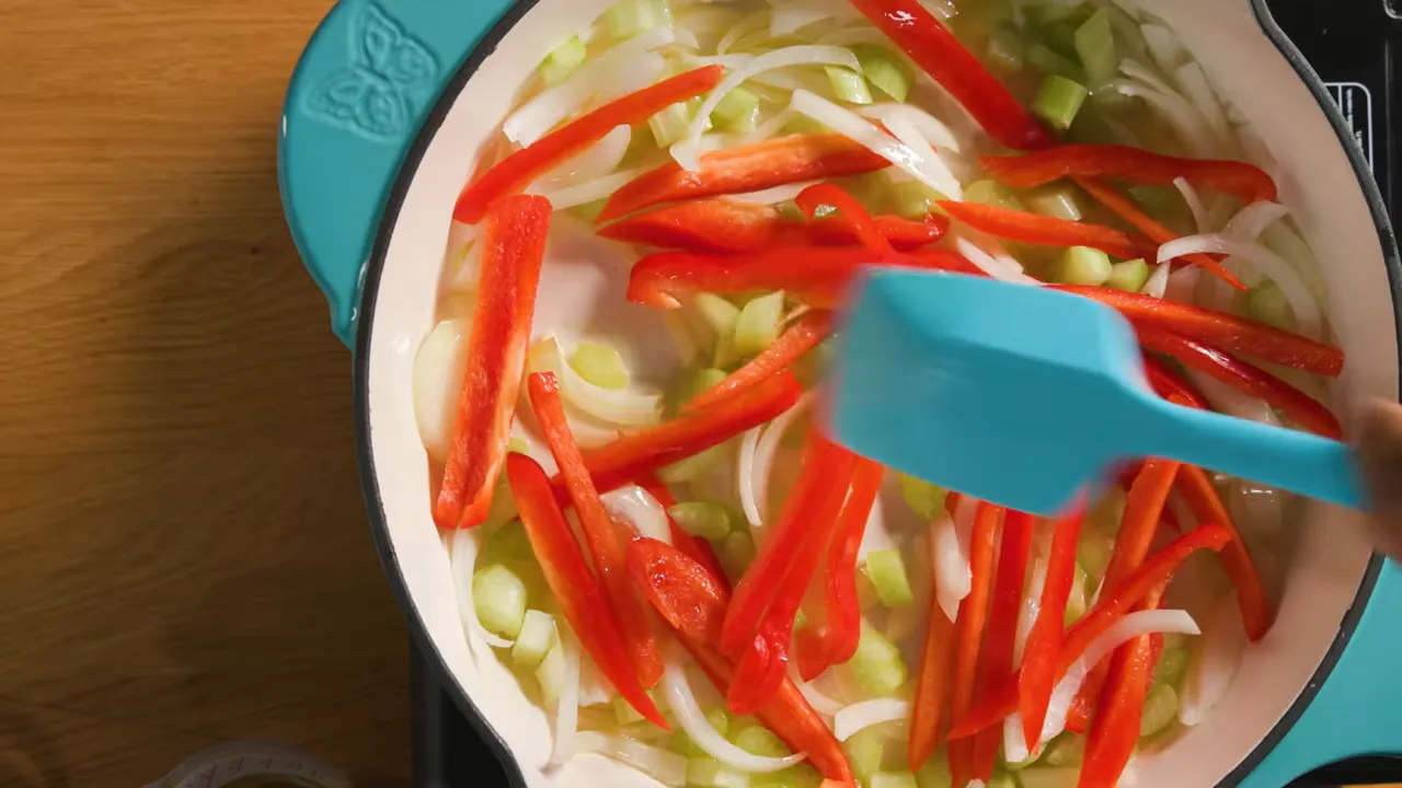 Close overhead view of onions, celery, and red bell pepper cooking in a Dutch oven with a blue spatula