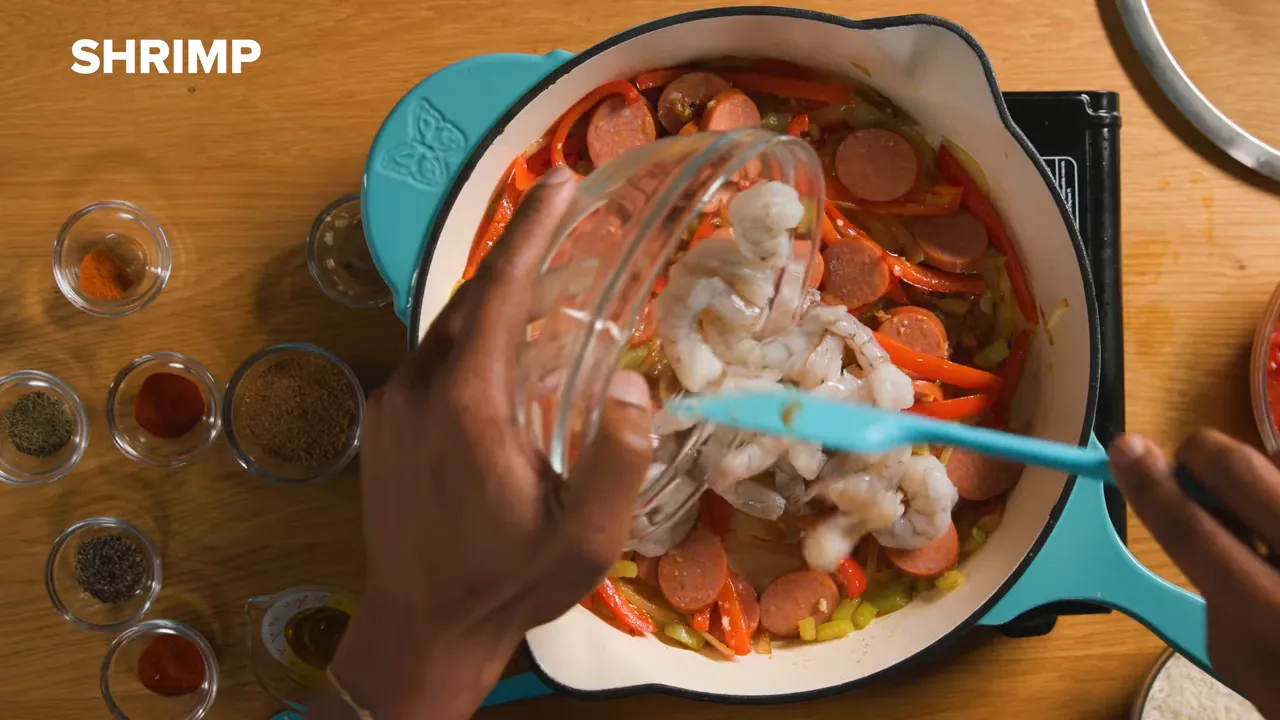 Overhead shot of shrimp being poured from a glass bowl into a Dutch oven with andouille, red pepper and celery; spices in small glass bowls line the counter.