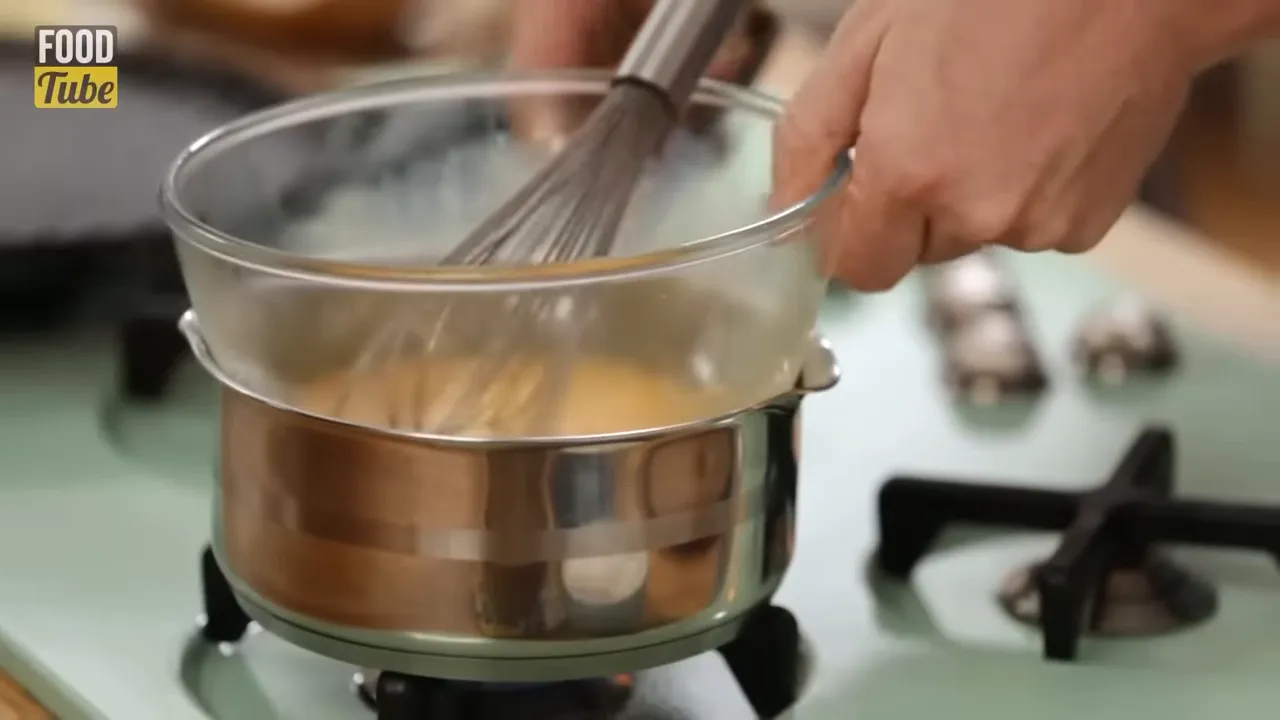 Close-up of a whisk stirring eggs in a glass bowl set over a pot of simmering water on the stove