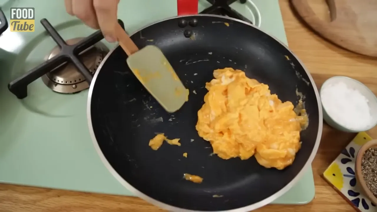 Top-down view of folded diner-style scrambled eggs in a nonstick pan with a spatula and small bowls of seasoning on the side
