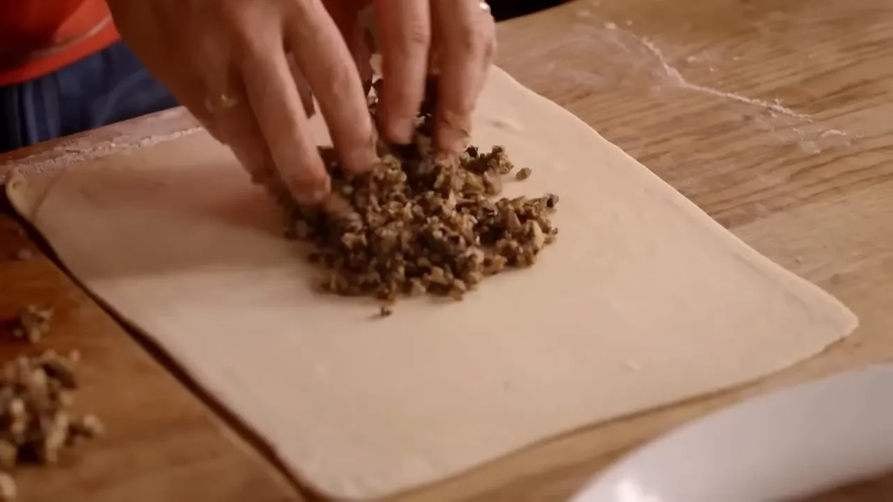 Hands placing mushroom and chicken liver mixture onto a sheet of puff pastry