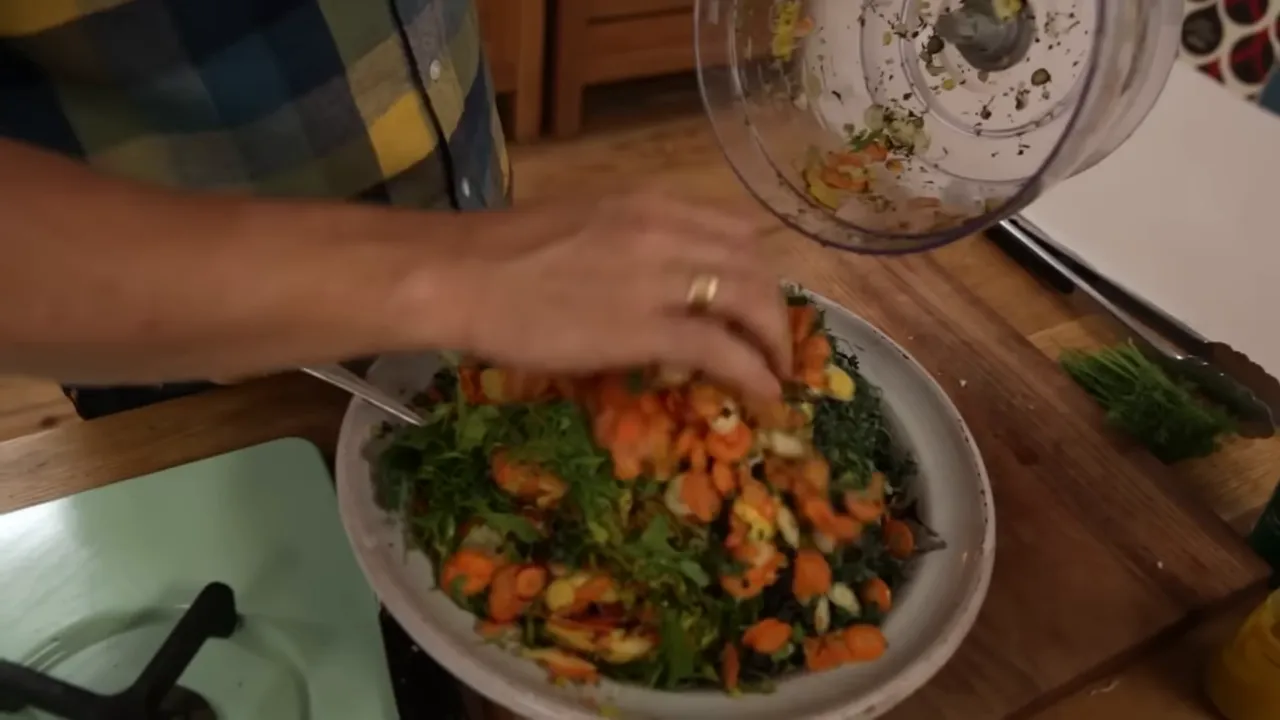 Pouring shredded carrots and mixed vegetables from a food processor into a bowl of greens for winter slaw
