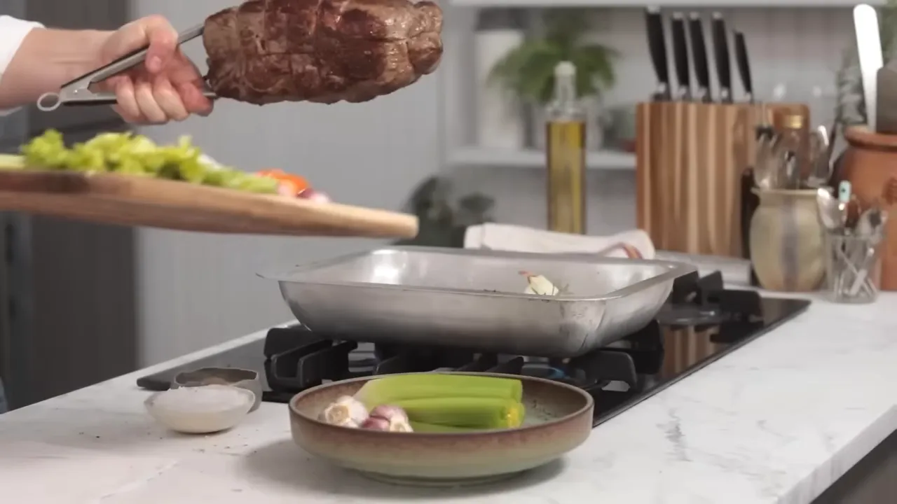Seared roast being lifted above a roasting tray filled with celery, garlic and onions ready to be placed in the oven.