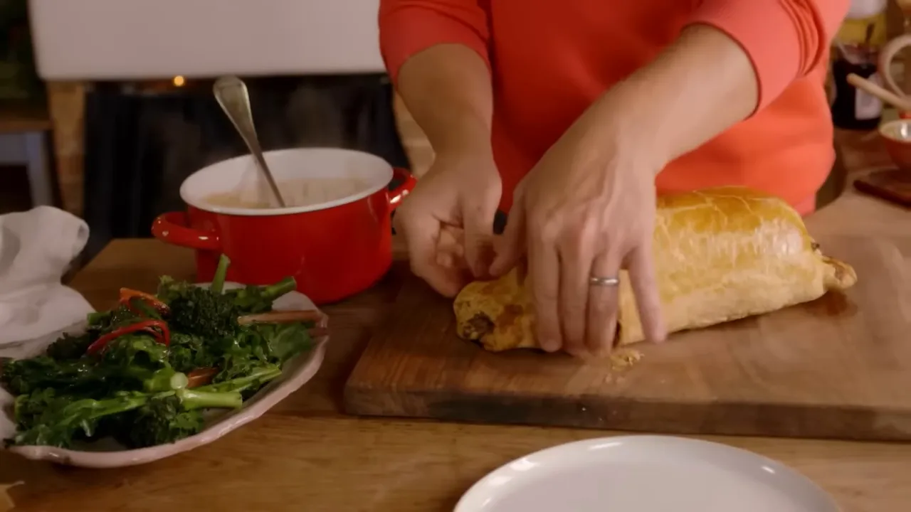 Beef Wellington on a wooden board being handled and prepared for slicing with greens and a pot of sauce beside it