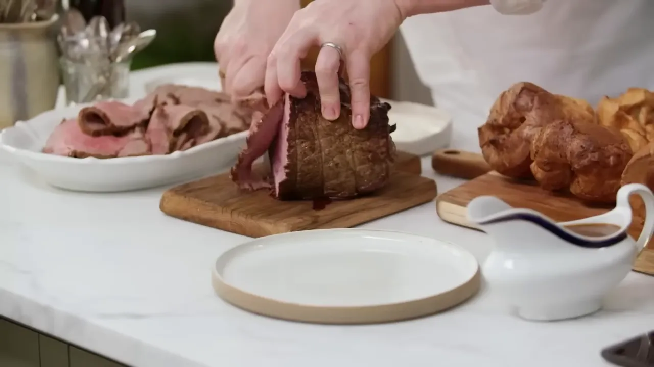 hands carving roast beef on a cutting board with Yorkshire puddings and a gravy boat on the counter