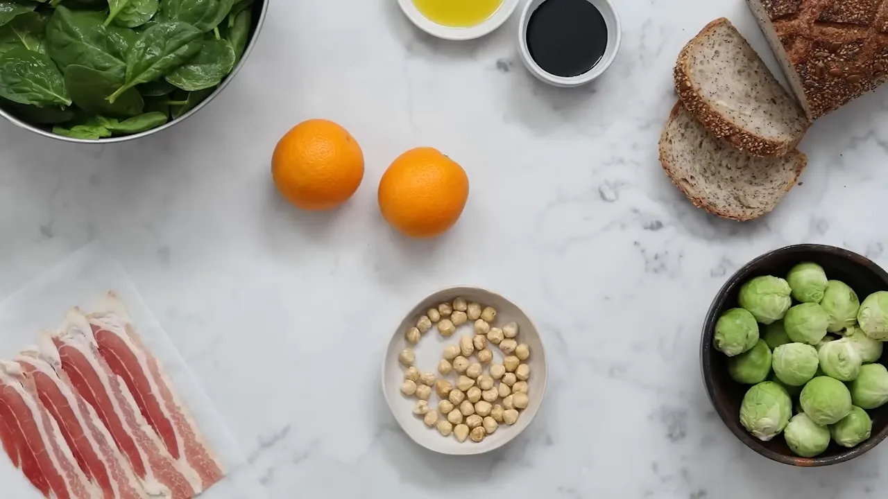 Overhead flatlay of spinach, two oranges, a bowl of hazelnuts, wholemeal seeded bread and a bowl of Brussels sprouts on a marble surface