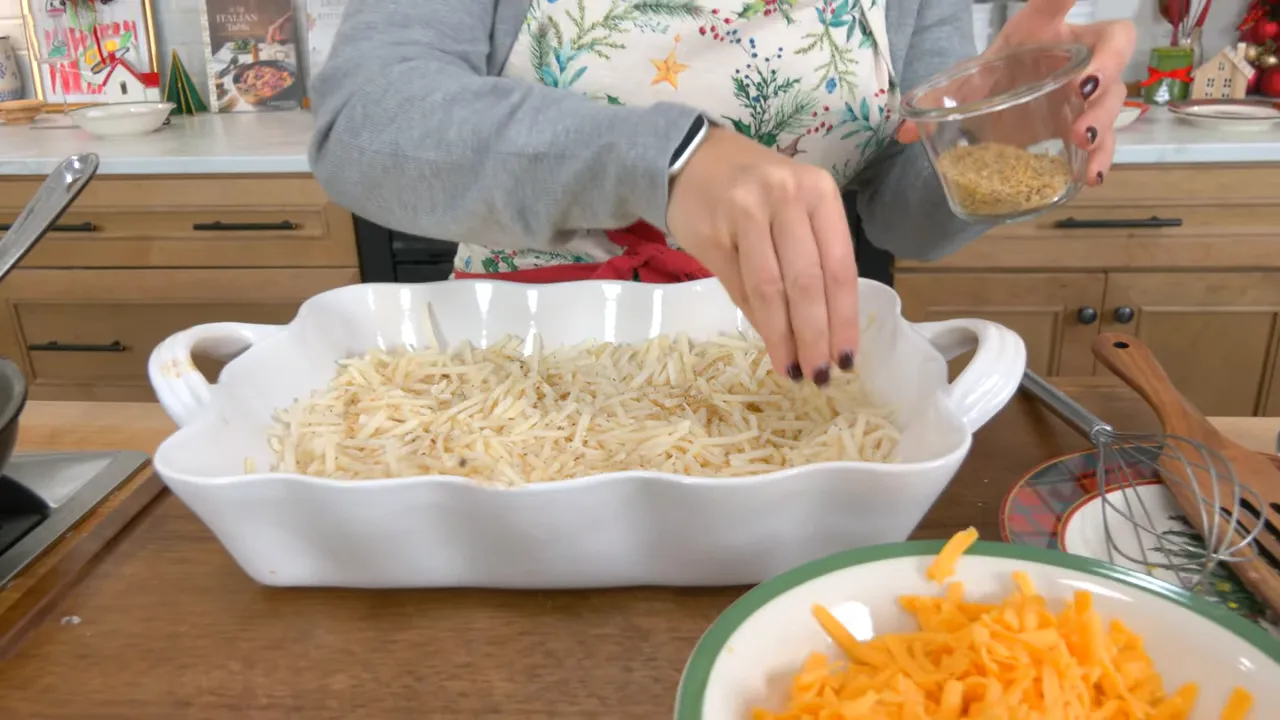 Hand sprinkling garlic-and-herb seasoning from a small glass jar over shredded hash browns in a white casserole dish, bowl of cheddar visible.