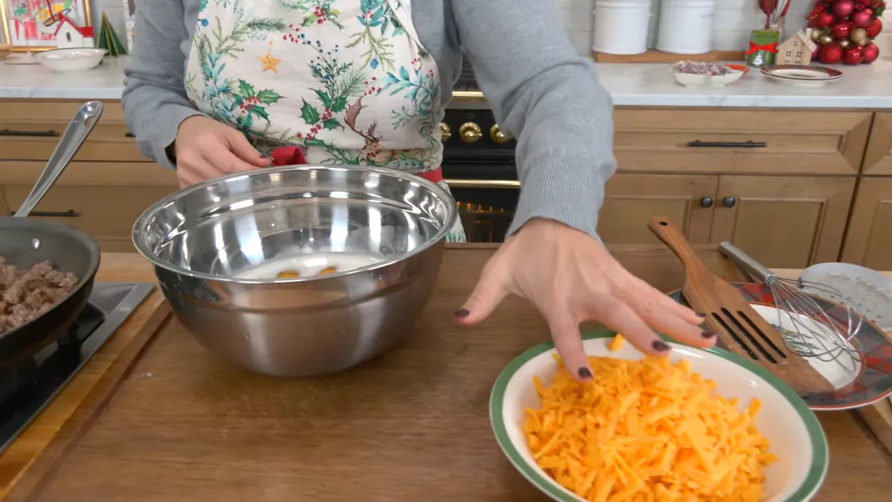 Mixing bowl with eggs and milk next to a bowl of freshly grated cheddar and a skillet of cooked sausage on the counter.