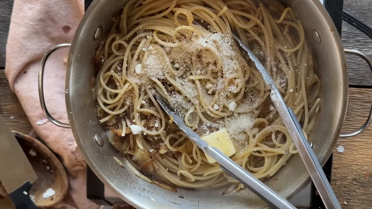 Top-down view of spaghetti being tossed in a pot with caramelized onions, a pat of butter and grated parmesan