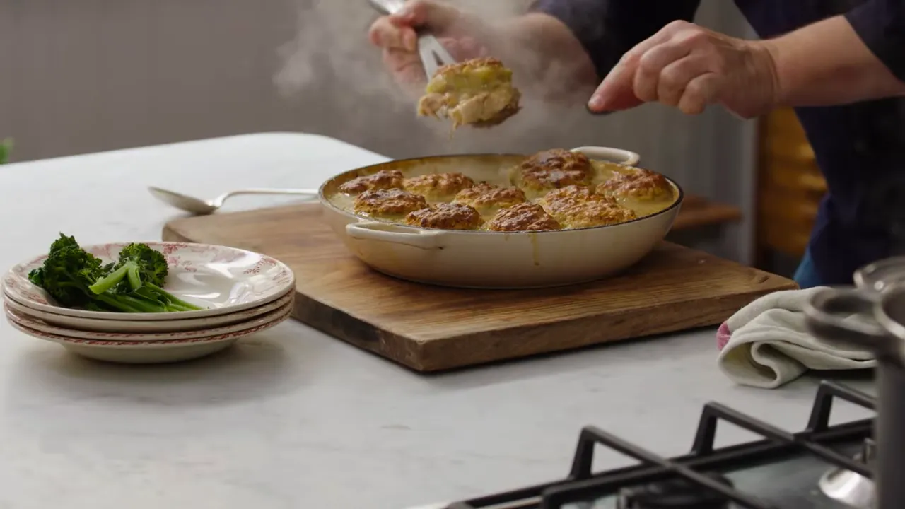 Skillet of turkey stew with golden biscuit dumplings on a wooden board with stacked plates and greens nearby