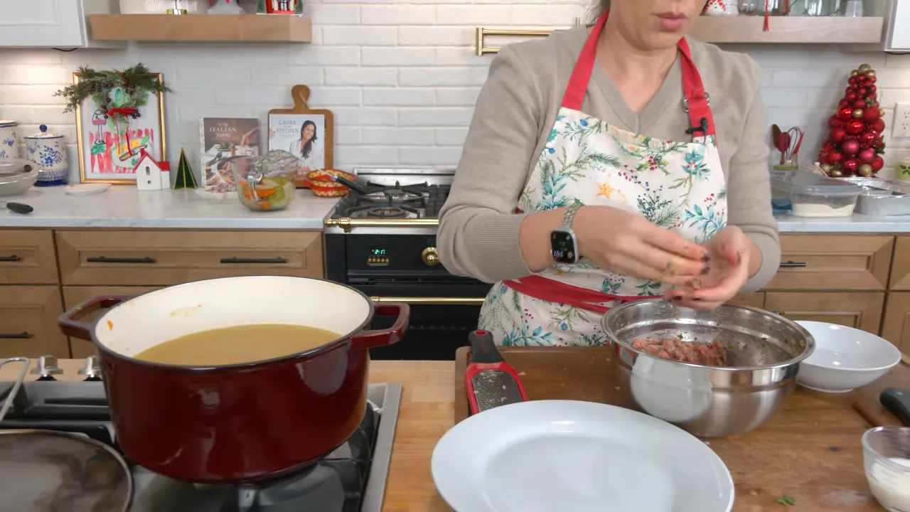 Red Dutch oven of clear chicken broth next to hands shaping mini meatballs in a metal bowl on a kitchen counter.