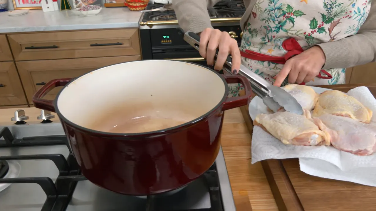 red Dutch oven on the stove with a hand using tongs to pick up bone-in, skin-on chicken thighs from a plate beside the pot