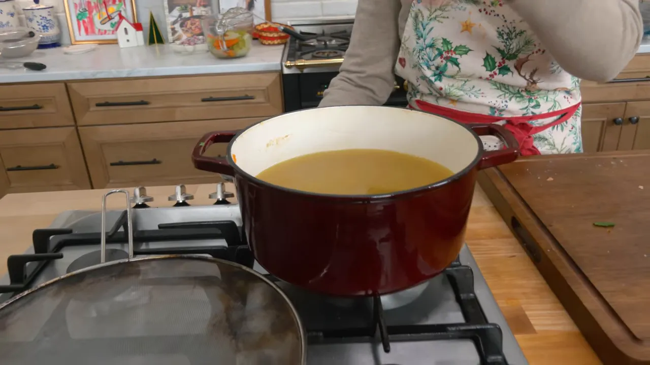 Red Dutch oven on the stovetop filled with clear homemade chicken broth with kitchen counter and tools in the background.