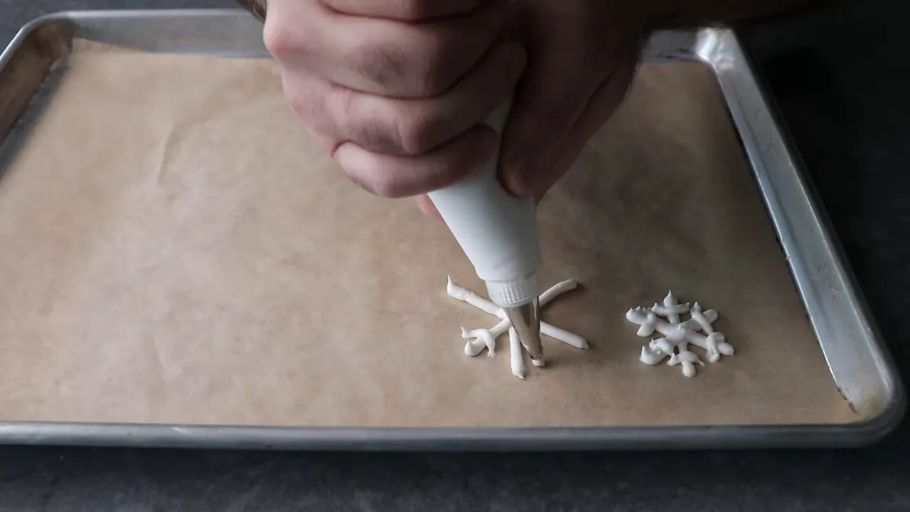 Hand-held piping bag adding finishing touches to a royal icing snowflake on parchment next to a small pile of finished snowflakes