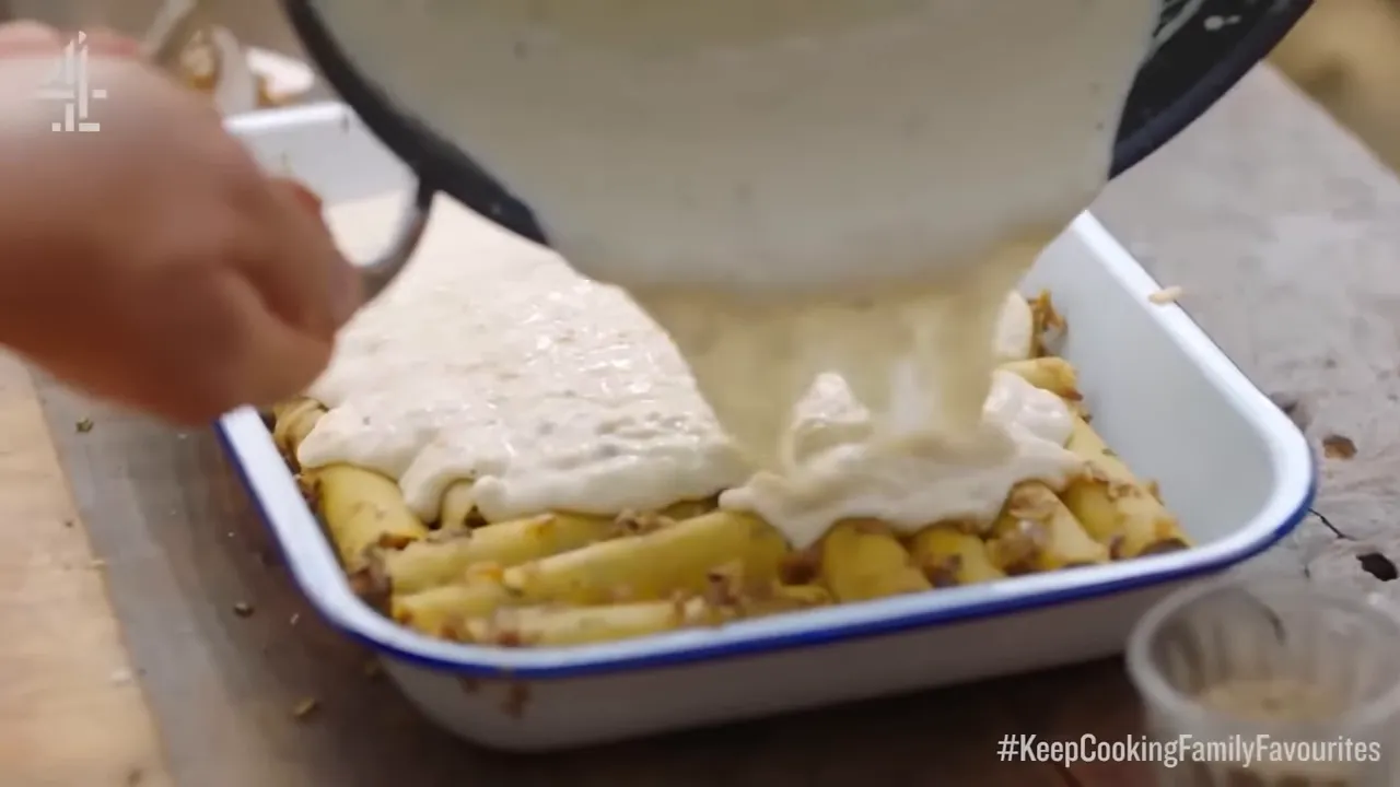 Hand tipping a pan to pour white sauce over mushroom-stuffed cannelloni in an enamel dish