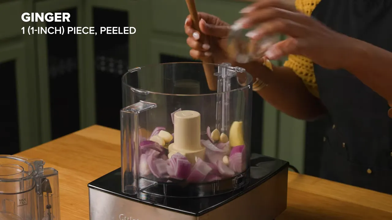 Food processor container with chopped onions, garlic and a piece of ginger being added.