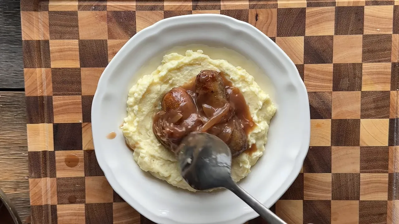 Overhead shot of bangers and mash with a spoon pouring onion gravy over the sausages on a white plate.