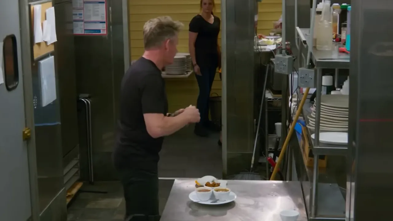 Kitchen pass with plated food, stacked dishes, cleaning tools and staff opening a doorway.