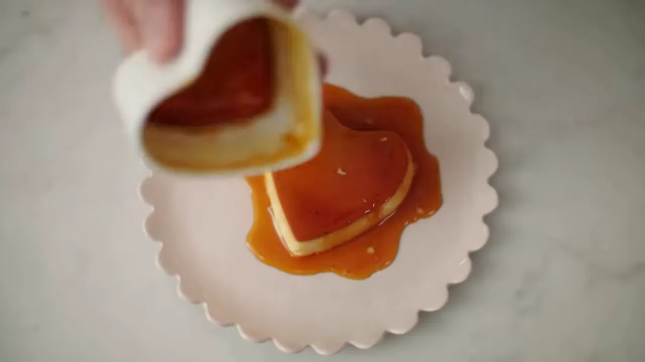 Top-down of a heart-shaped creme caramel on a pink scalloped plate with caramel sauce being poured from the mould