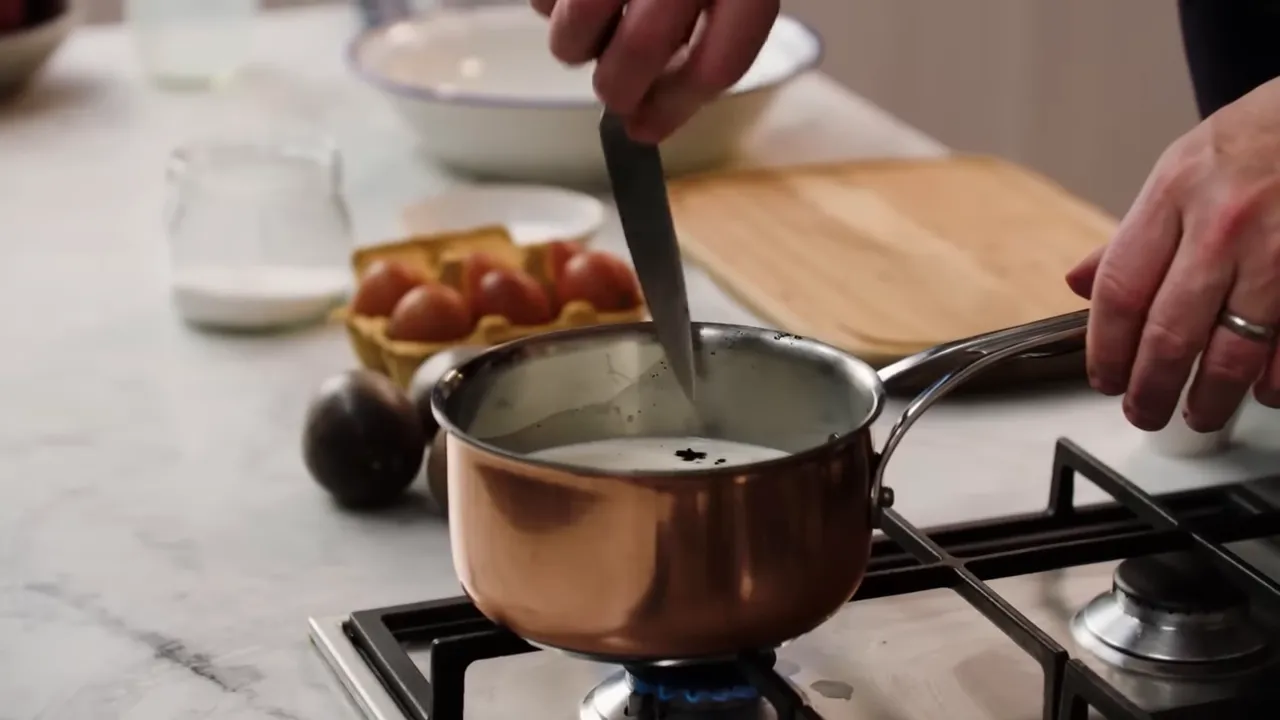 hand stirring vanilla-infused milk in a copper saucepan with eggs and prep items in the background