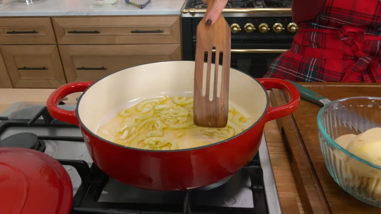 Close-up of a red enamel Dutch oven with thinly sliced onions cooking in olive oil and a wooden spatula