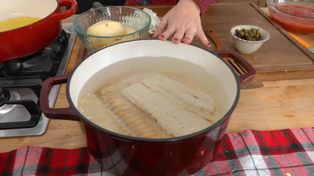 Large red enameled pot on a counter with baccala fillets submerged in water; potatoes, olives and a bowl of tomatoes visible nearby.