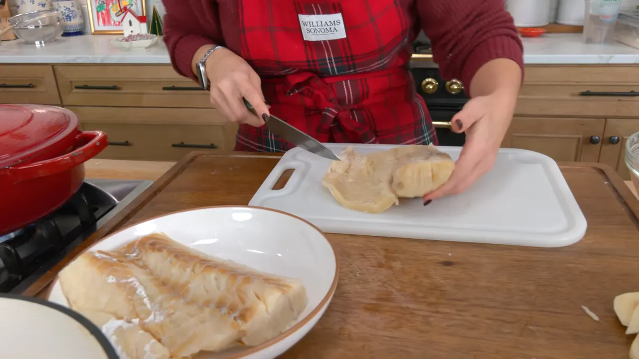 Hands slicing rehydrated baccala (salted cod) on a cutting board with a plate of fish and a red pot on the stove nearby