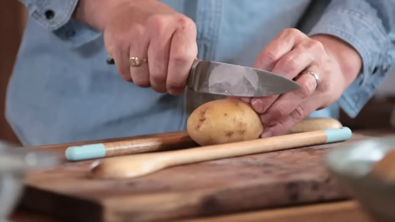 Hands slicing a potato between two wooden spoons to create even Hasselback cuts, showing knife technique