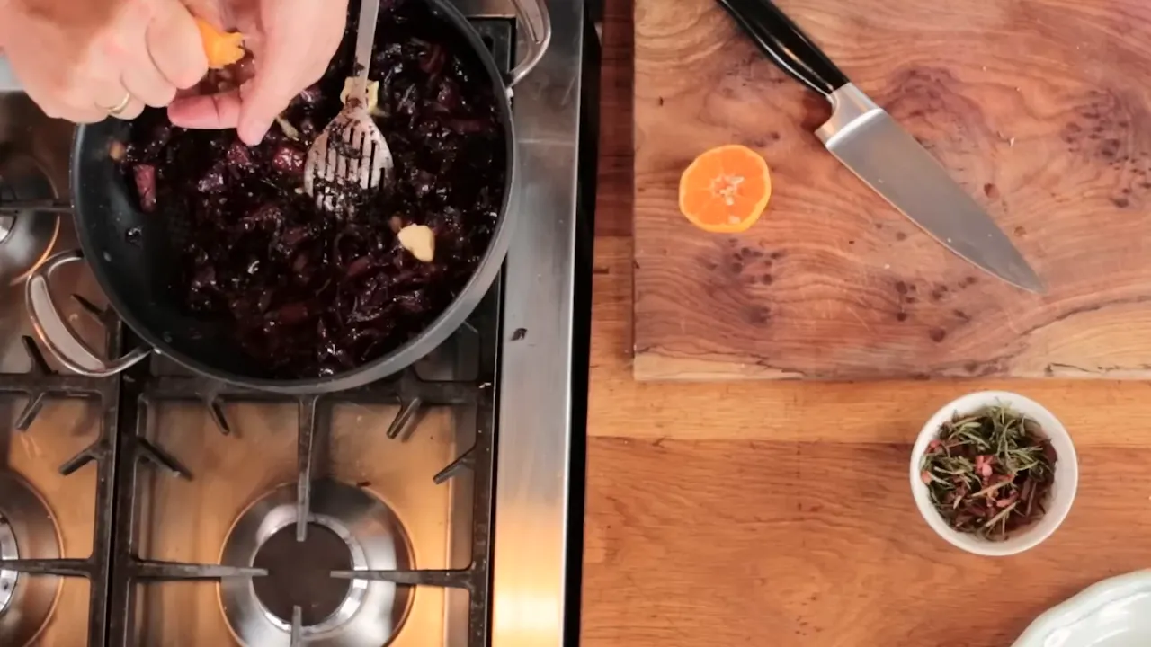top-down of hands squeezing a clementine over a pan of braised red cabbage with butter and a small bowl of herbs on the cutting board