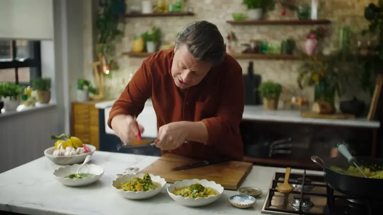 Cook leaning over bowls of vegetable soup on a countertop, grating or adding a garnish over them with a pan simmering on the hob