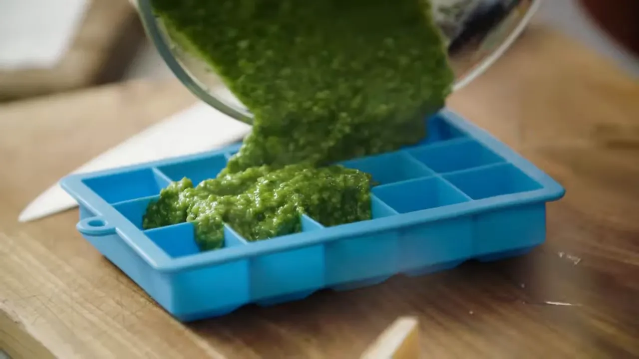close-up pouring fresh green pesto into a blue silicone ice cube tray ready for freezing