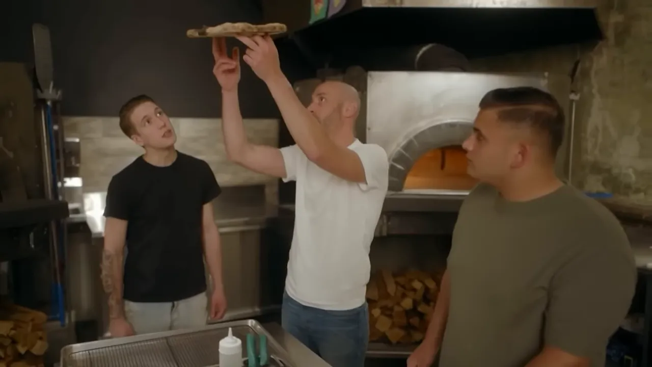 Chef holding a pizza up to check crust color and doneness in front of a wood‑fired oven.