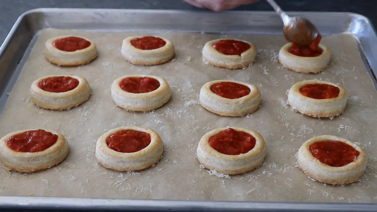 Tray of puff pastry pizza poppers on parchment being filled with pizza sauce using a spoon.