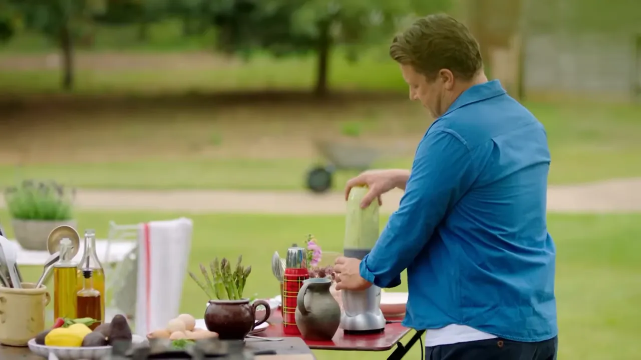 Host using a blender on an outdoor table to make an avocado hollandaise with asparagus, eggs and other ingredients nearby.