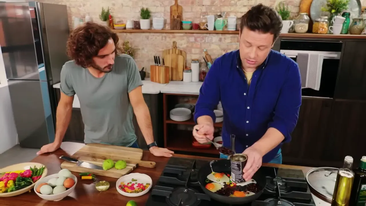 Wide kitchen shot showing eggs frying in a pan with black beans, avocado halves on a cutting board and bowls of ingredients on the counter.