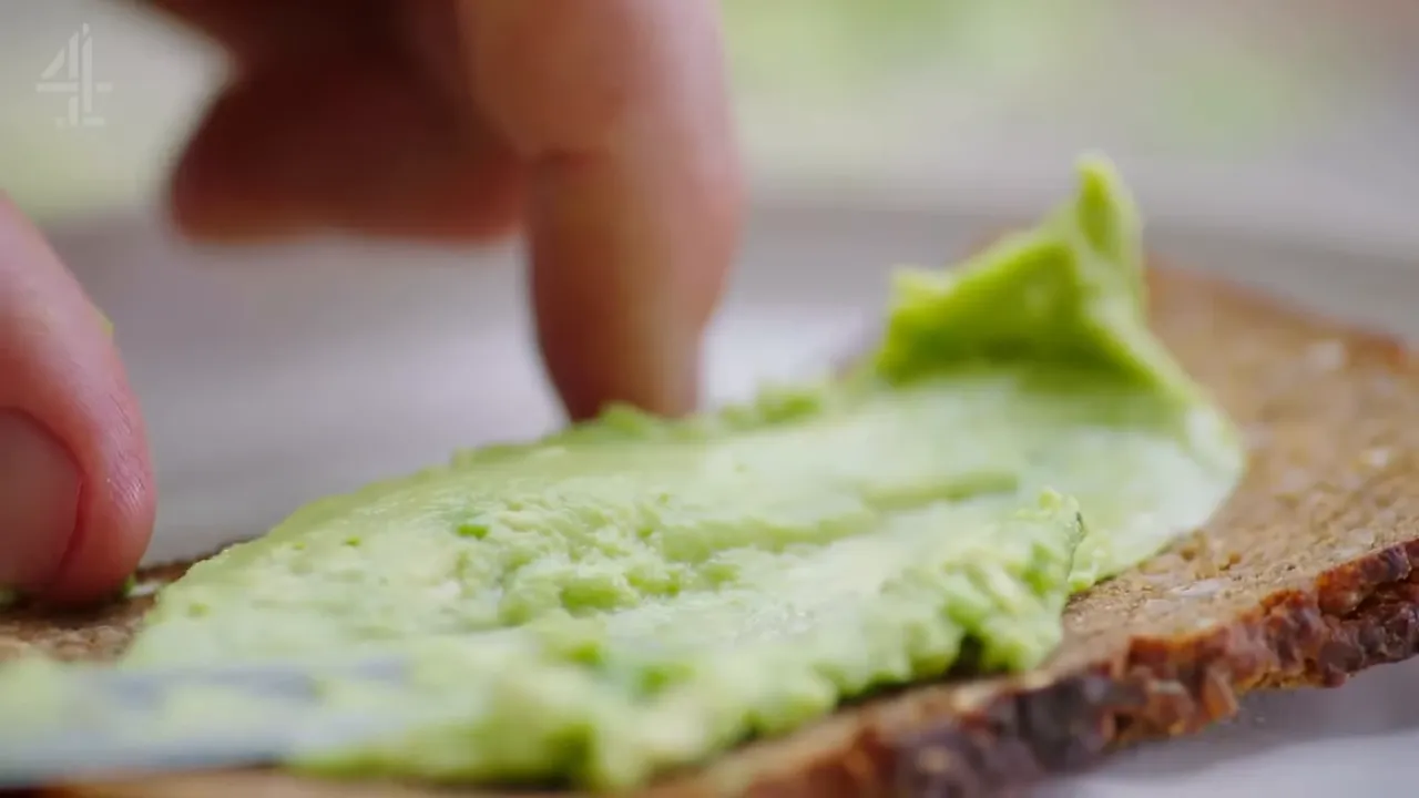 mashed avocado being spread onto a slice of rye bread with a finger and knife