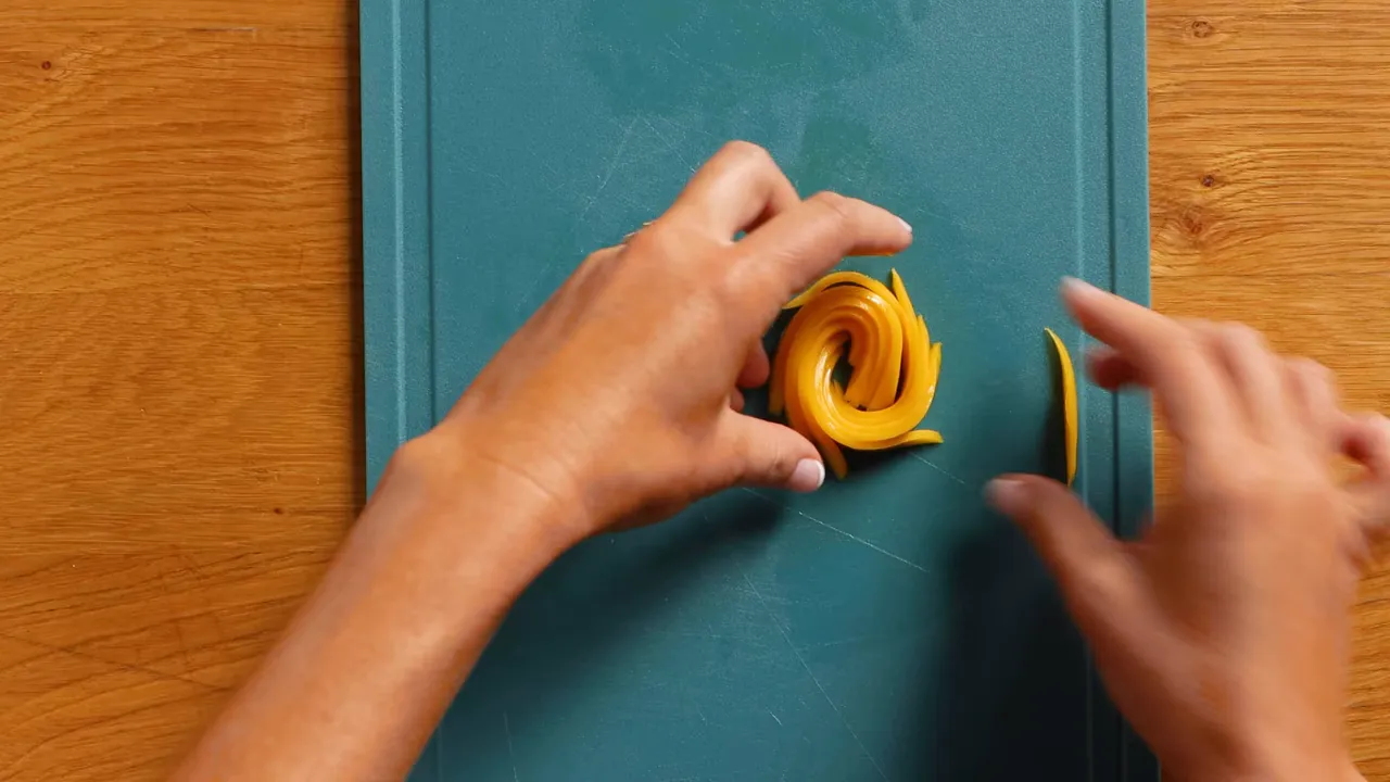 Top-down clear image of a completed mango rose made from thin slices on a cutting board with hands nearby