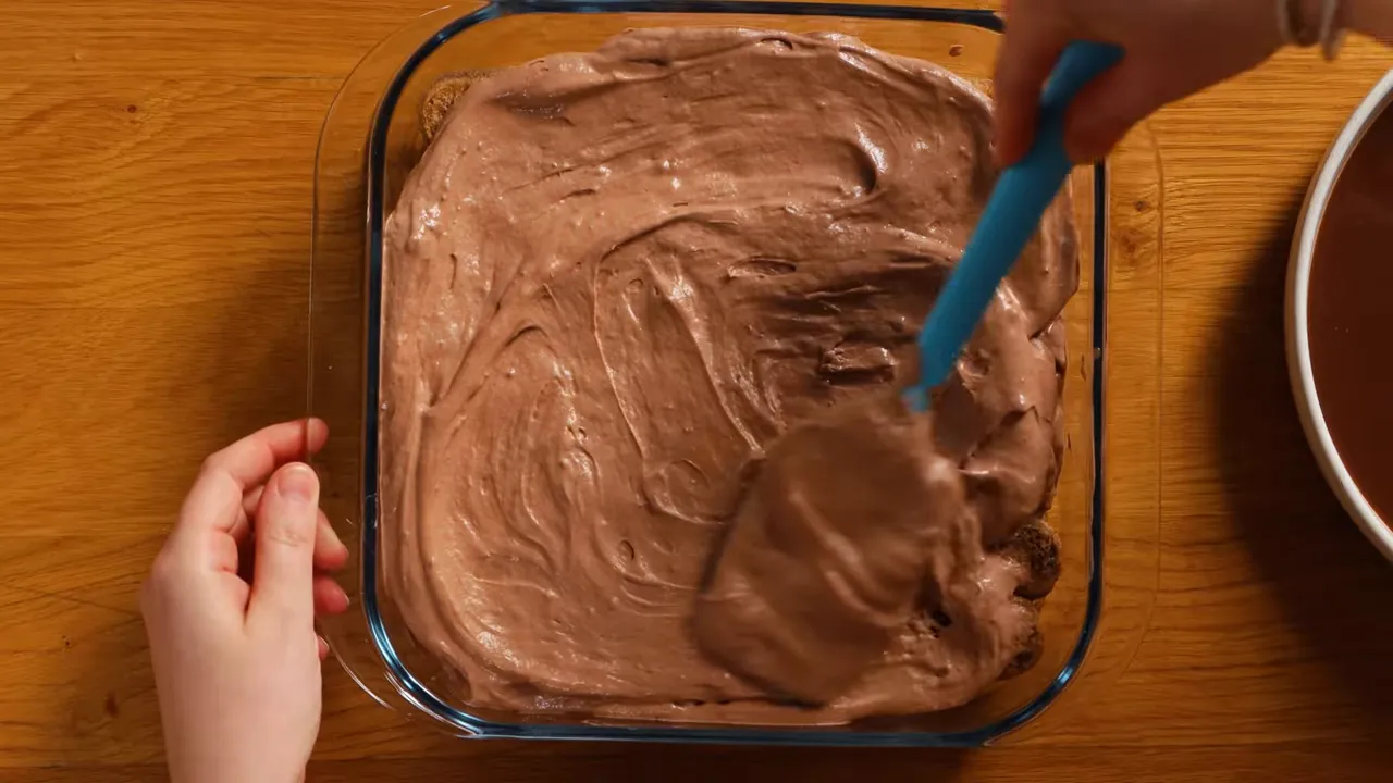 Top-down view of a spatula smoothing chocolate tiramisu filling over ladyfingers in a glass pan