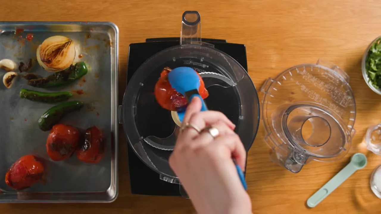 Top-down view of a roasted tomato being spooned into a food processor with a tray of charred vegetables beside it.