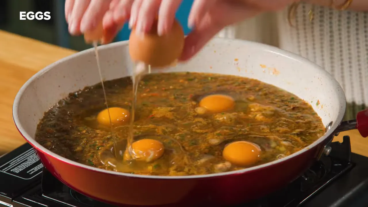 Close-up of eggs being cracked into a red pan of simmering tomato-chile salsa