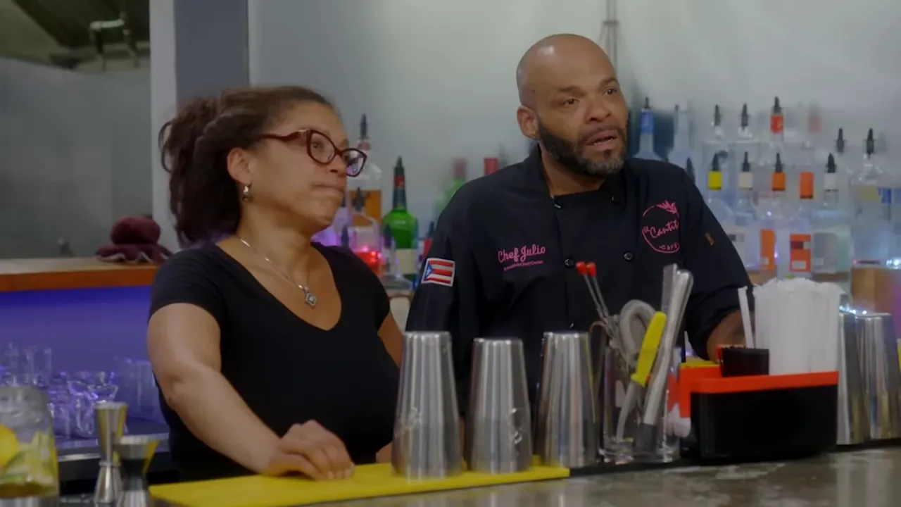 Two restaurant owners standing behind the counter with bar tools and metal cups, listening and engaged in discussion.