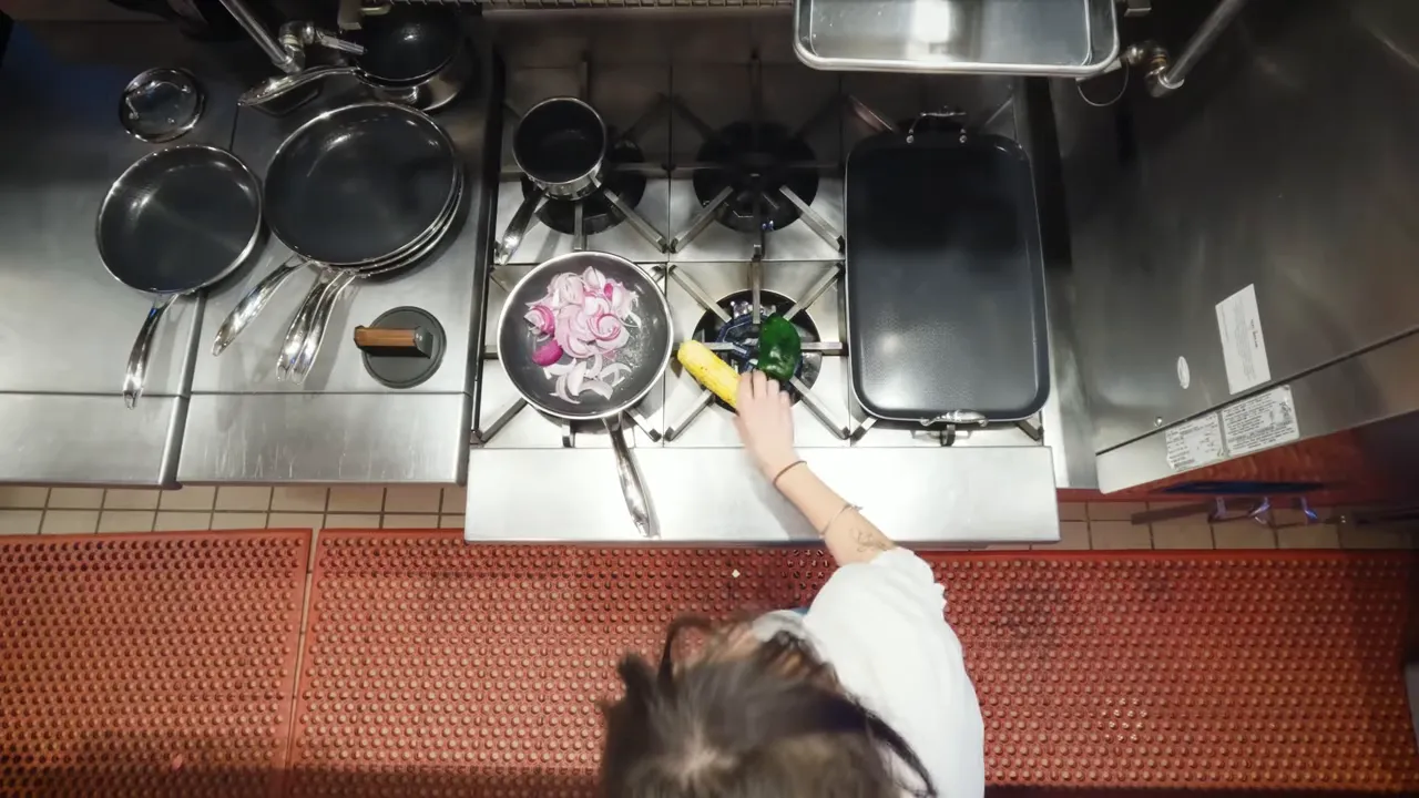Overhead shot of a pan with sliced red onions and a corn cob being charred on a commercial stove