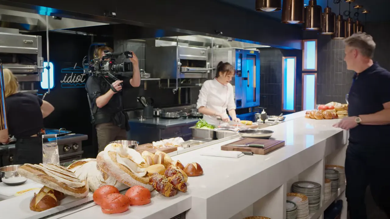 wide kitchen pass with assorted breads in the foreground, a chef prepping behind the counter and another person watching