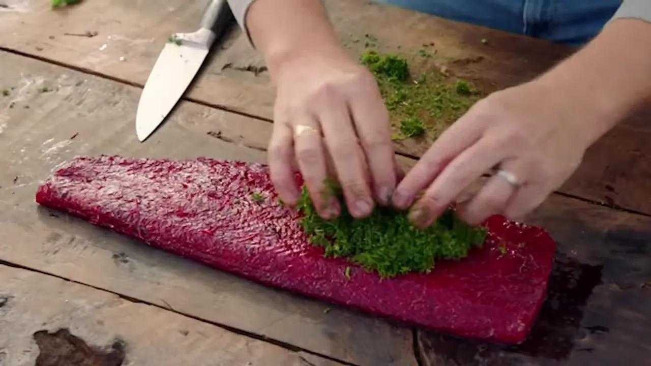 Hands pressing chopped dill and herbs onto a bright beetroot-cured salmon fillet on a wooden board