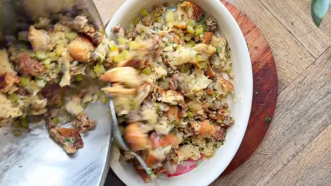 Overhead view of stuffing mixture of torn bread, sausage, celery and herbs being scraped into a baking dish