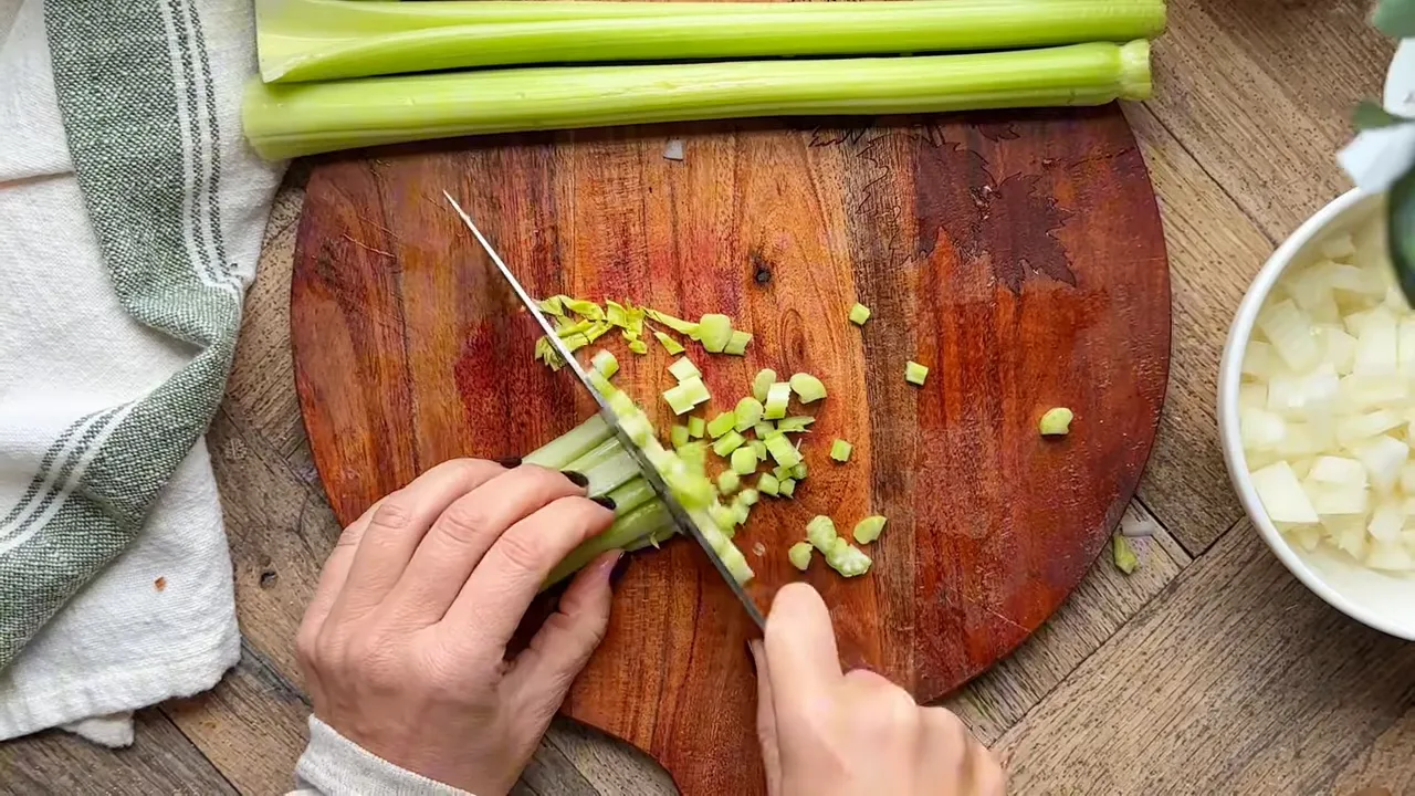Overhead shot of hands chopping celery on a wooden cutting board with whole stalks at the top and a bowl of diced onion to the right